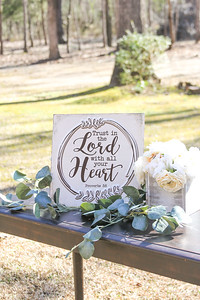 Welcome table at an outdoor wedding with greenery, white flowers and a Bible verse sign, "Trust in the Lord"