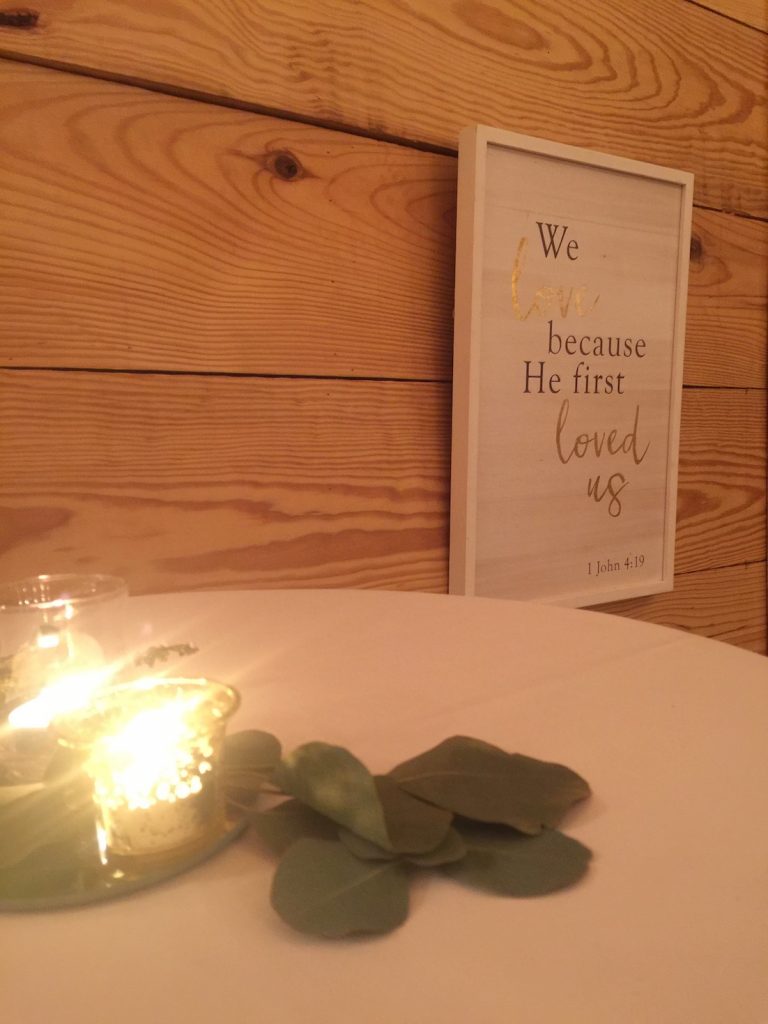 Candles on a white table cloth with a white wedding Bible verse sign, "We love because He first loved us" hangin on a pine wood wall